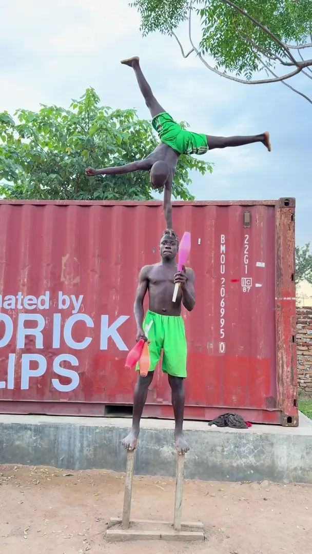A Clon Circus Uganda acrobat balances above a partner who is juggling clubs outdoors.